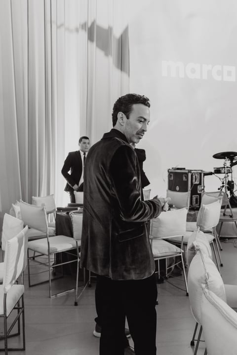 Black and white photo of a man in a leather jacket standing in a modern office space with a drum kit, another man in a suit visible in the background