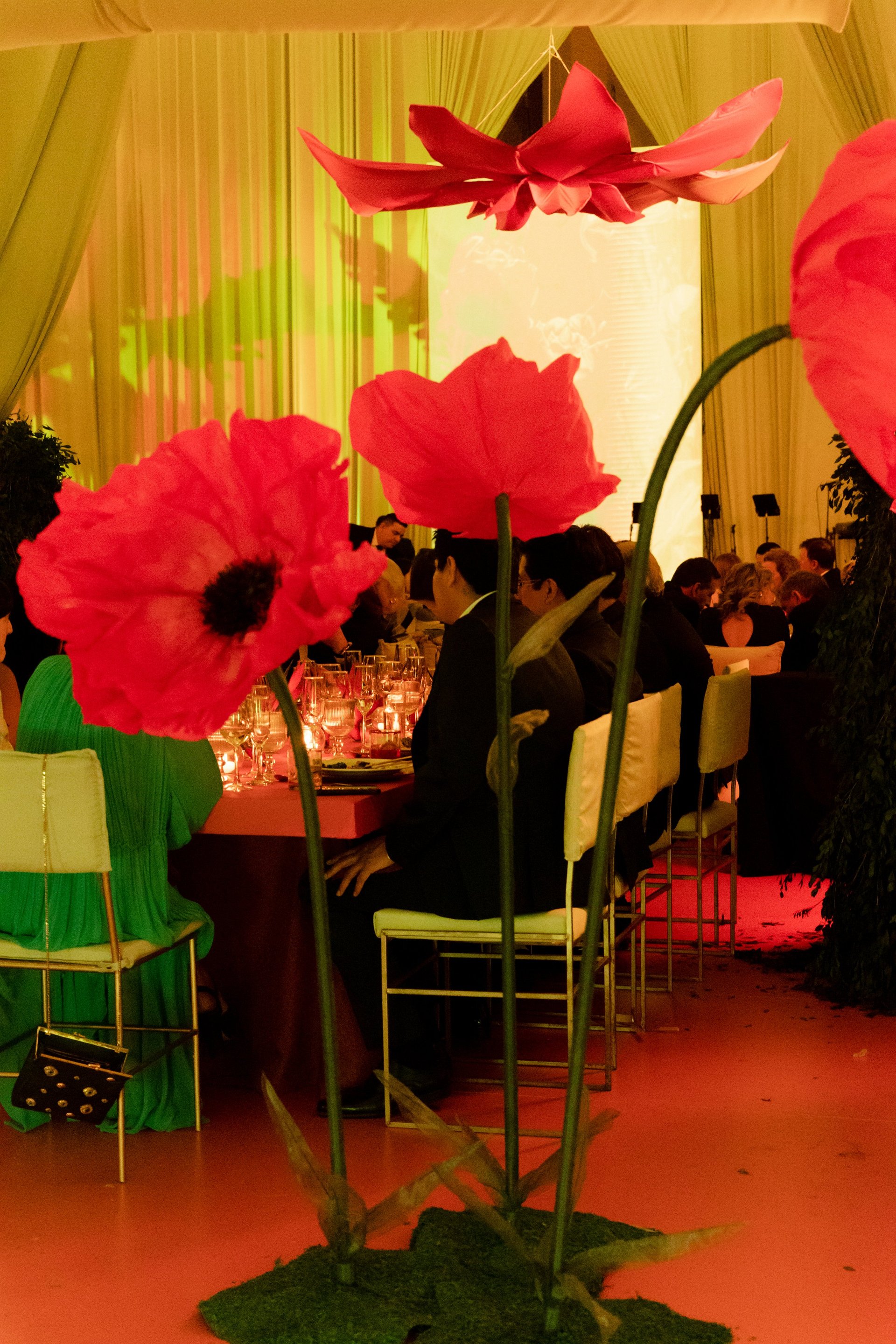 Giant red poppy flowers with black centers displayed indoors at an event with gold curtains and people in the background