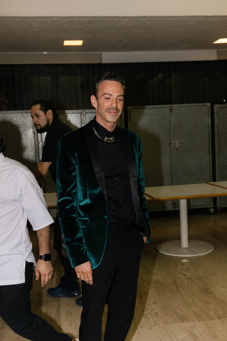 Man in teal velvet blazer and black shirt with chain necklace standing in modern indoor venue