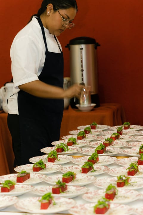 Catering worker in black apron preparing appetizer plates with strawberries and greens on an orange background